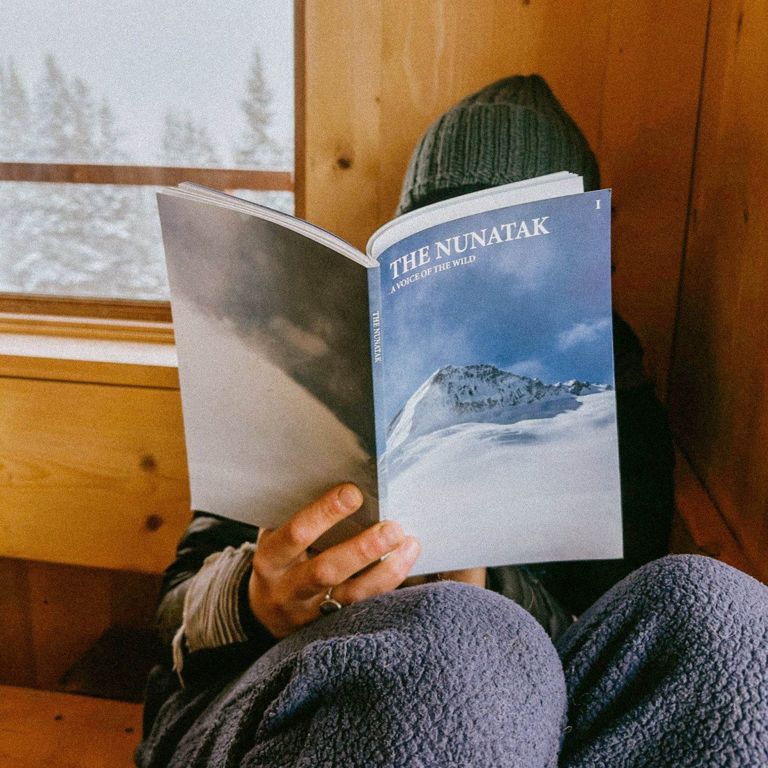 Person reading a book titled 'The Nunatak' in a cozy cabin setting.